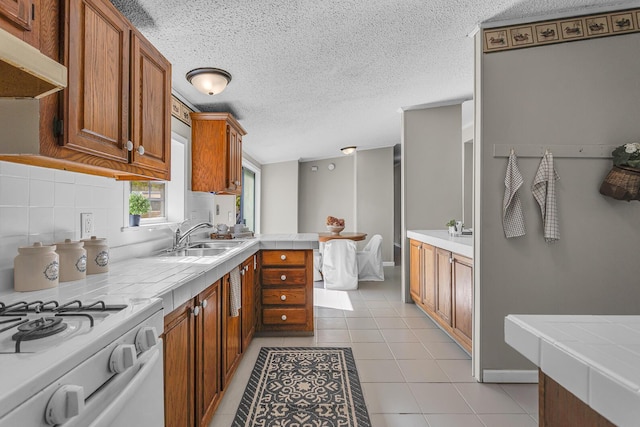 kitchen with brown cabinetry, white gas range, under cabinet range hood, tasteful backsplash, and a sink