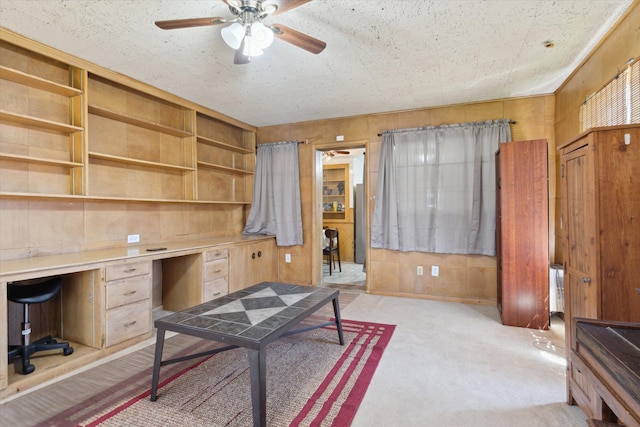 home office featuring wooden walls, ceiling fan, light carpet, and built in desk