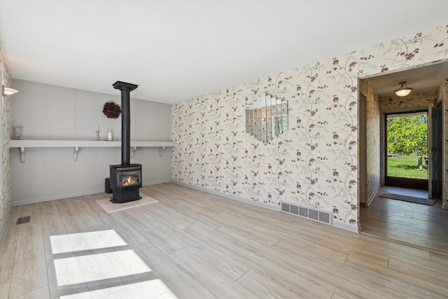 unfurnished living room with visible vents, a wood stove, wood finished floors, baseboards, and wallpapered walls