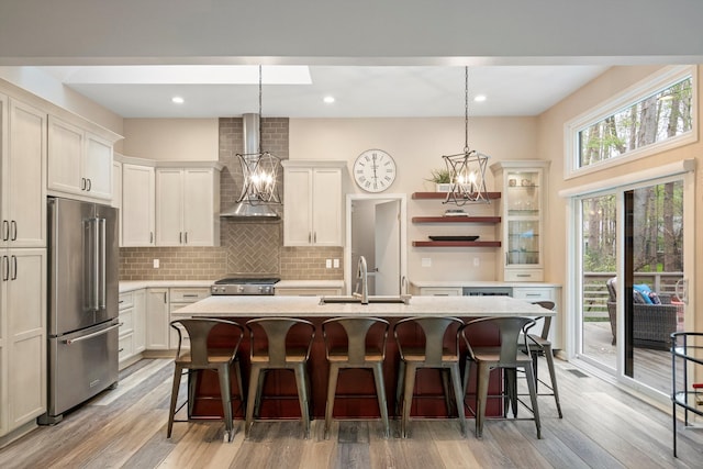 kitchen with open shelves, high quality fridge, a sink, wall chimney exhaust hood, and an island with sink