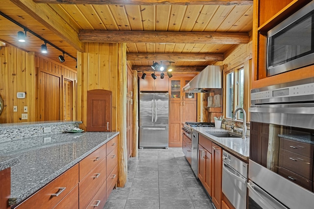 kitchen featuring exhaust hood, appliances with stainless steel finishes, a sink, wooden walls, and dark stone counters