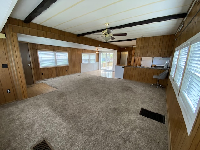 unfurnished living room with visible vents, wood walls, beam ceiling, a ceiling fan, and carpet