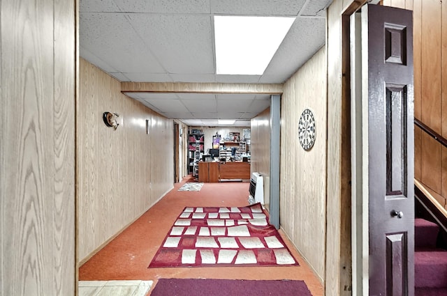 hall with carpet floors, wood walls, a paneled ceiling, and heating unit