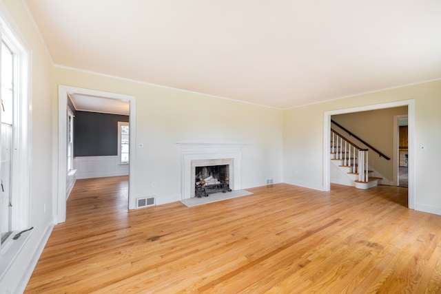 unfurnished living room featuring stairs, a fireplace with flush hearth, light wood-type flooring, visible vents, and ornamental molding