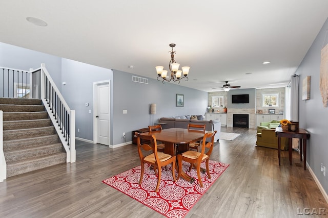 dining area featuring visible vents, a large fireplace, wood finished floors, ceiling fan with notable chandelier, and stairs