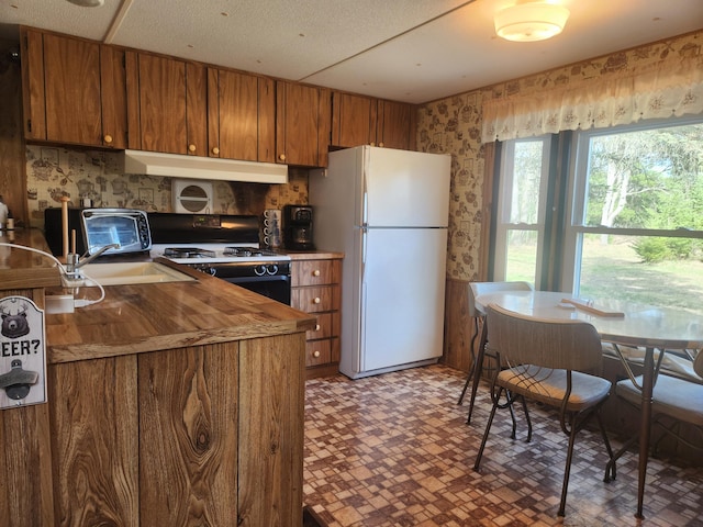 1837 9 Mile Rd, Sears, MI 49679 - kitchen featuring freestanding refrigerator, black gas range oven, under cabinet range hood, brown cabinetry, and wallpapered walls