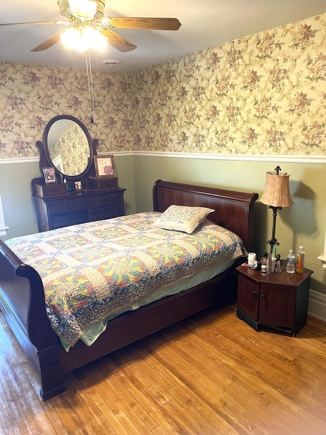 bedroom with wood finished floors, ceiling fan, a wainscoted wall, and wallpapered walls
