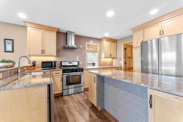 kitchen featuring wall chimney range hood, appliances with stainless steel finishes, a sink, light wood-style floors, and light brown cabinets