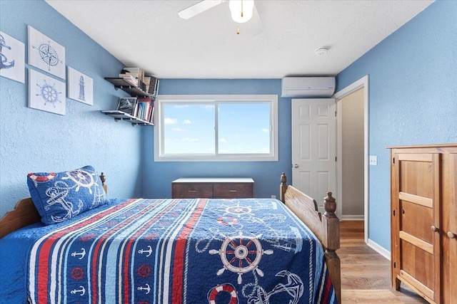 bedroom with a textured wall, ceiling fan, baseboards, a wall unit AC, and wood finished floors