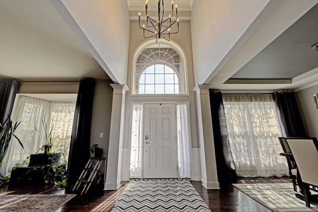 entryway featuring decorative columns, a towering ceiling, crown molding, a chandelier, and dark wood-type flooring