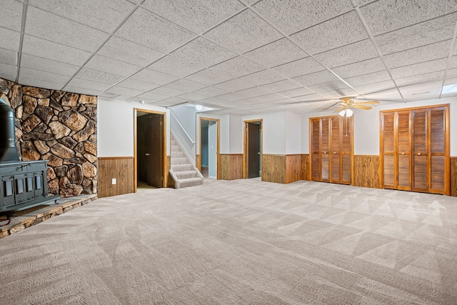 finished basement with a wood stove, wainscoting, a drop ceiling, and stairway