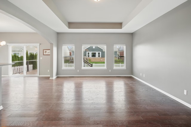 empty room with arched walkways, a tray ceiling, wood finished floors, and a healthy amount of sunlight