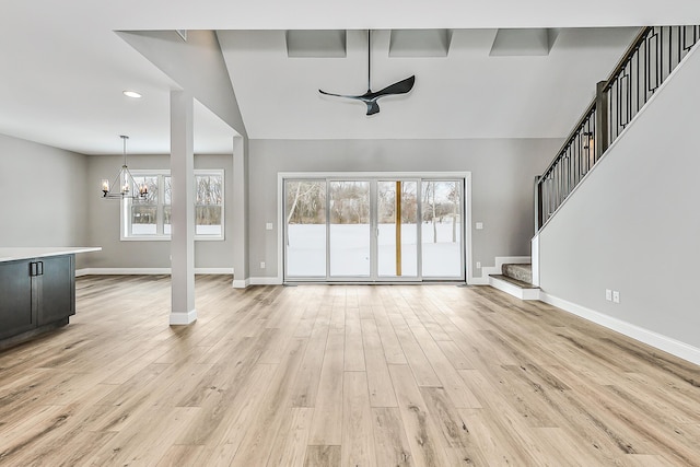 unfurnished living room featuring light wood finished floors, ceiling fan with notable chandelier, baseboards, vaulted ceiling, and stairway