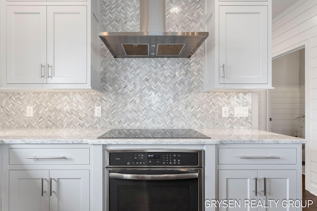 kitchen with wall chimney range hood, tasteful backsplash, light stone countertops, black electric stovetop, and stainless steel oven