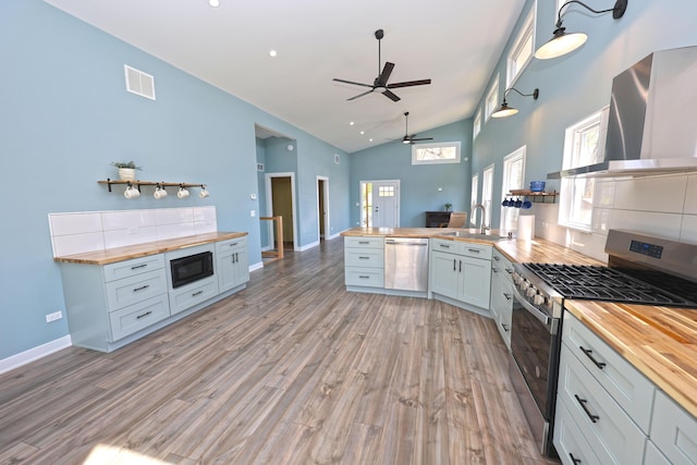 kitchen with appliances with stainless steel finishes, wood counters, wall chimney exhaust hood, a ceiling fan, and visible vents