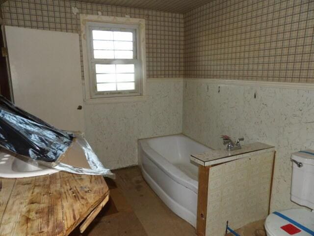 full bath featuring wainscoting, toilet, wallpapered walls, wood finished floors, and a garden tub