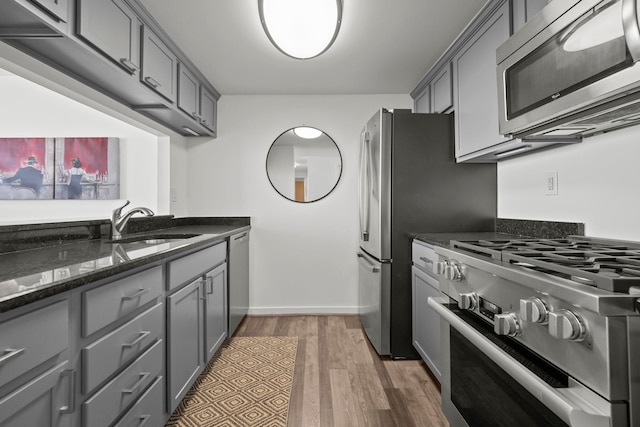 kitchen with stainless steel appliances, gray cabinetry, a sink, dark stone counters, and wood finished floors