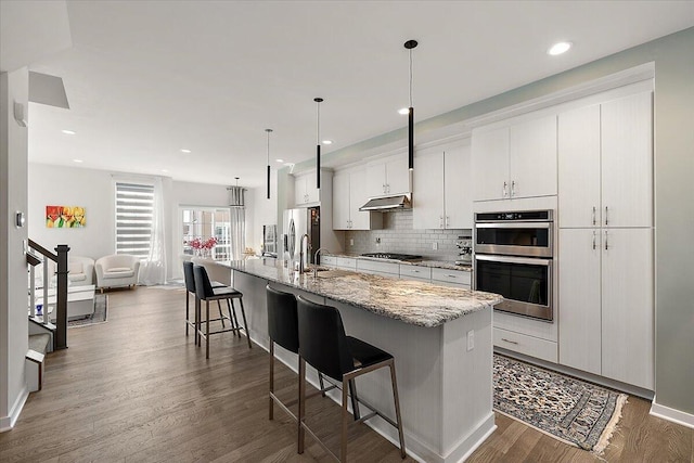kitchen featuring a breakfast bar area, under cabinet range hood, a large island with sink, dark wood finished floors, and appliances with stainless steel finishes