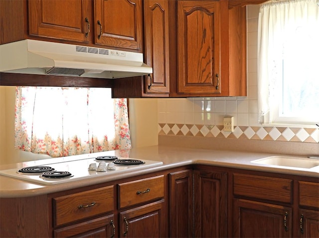 kitchen with brown cabinetry, white electric stovetop, light countertops, under cabinet range hood, and backsplash