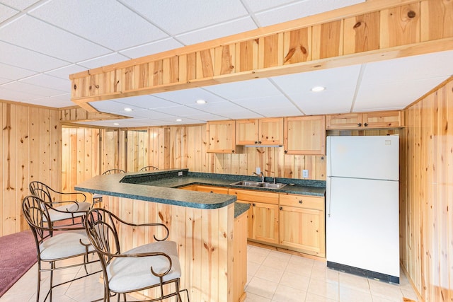 kitchen featuring dark countertops, a sink, freestanding refrigerator, wooden walls, and light brown cabinets