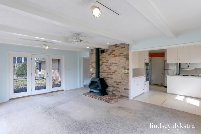 unfurnished living room featuring a wood stove, light carpet, beamed ceiling, ceiling fan, and french doors
