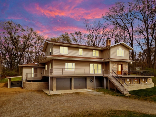 back of property at dusk with a deck, a chimney, stairs, dirt driveway, and an attached garage
