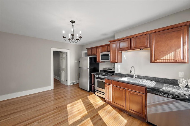 kitchen with brown cabinets, appliances with stainless steel finishes, an inviting chandelier, a sink, and light wood-type flooring