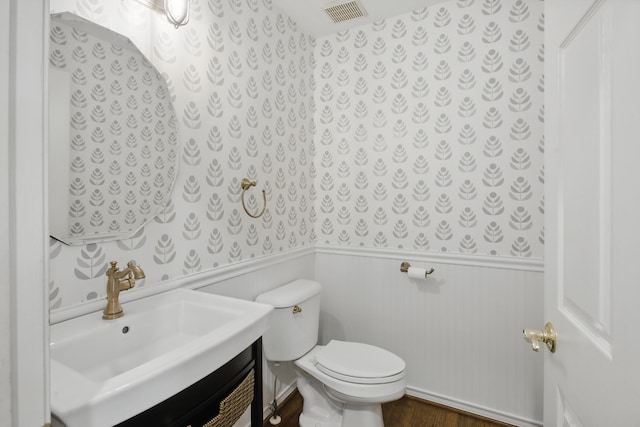 half bath with visible vents, vanity, wood finished floors, toilet, and a wainscoted wall