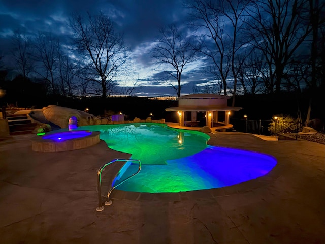 pool at night with fence, a pool with connected hot tub, a storage structure, a patio area, and an outdoor structure