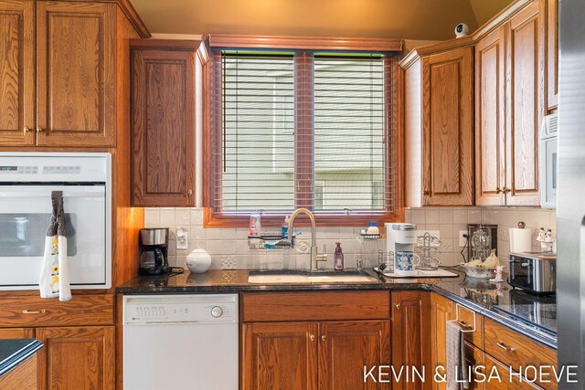 kitchen with brown cabinets, a sink, white appliances, dark stone countertops, and tasteful backsplash