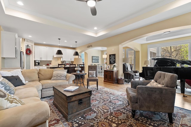 living room with arched walkways, ceiling fan, wood finished floors, and a raised ceiling