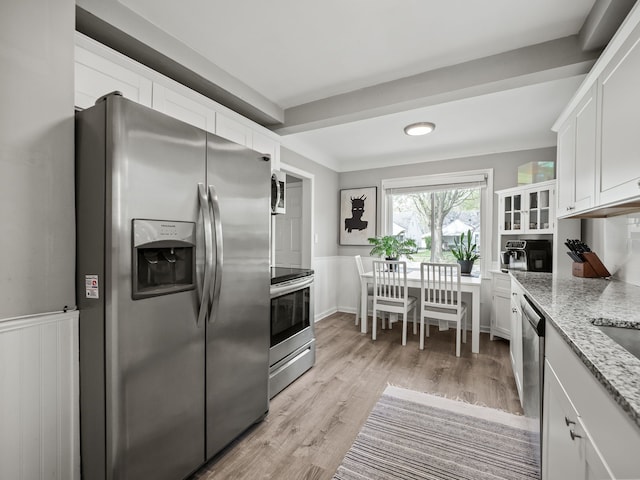 kitchen with white cabinets, appliances with stainless steel finishes, light stone counters, light wood finished floors, and a wainscoted wall