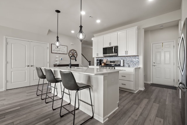 kitchen with a kitchen island with sink, backsplash, wood finished floors, stainless steel appliances, and white cabinetry