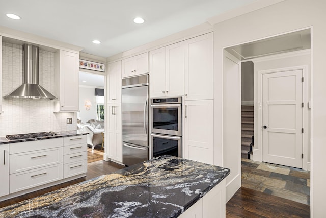 kitchen featuring wall chimney range hood, dark stone counters, white cabinets, backsplash, and stainless steel appliances