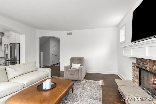 living room with a stone fireplace, visible vents, arched walkways, baseboards, and dark wood-style floors