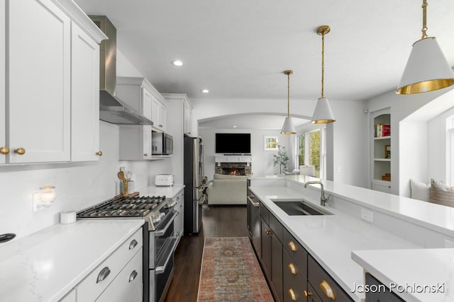 kitchen featuring open floor plan, a warm lit fireplace, wall chimney exhaust hood, a sink, and stainless steel appliances