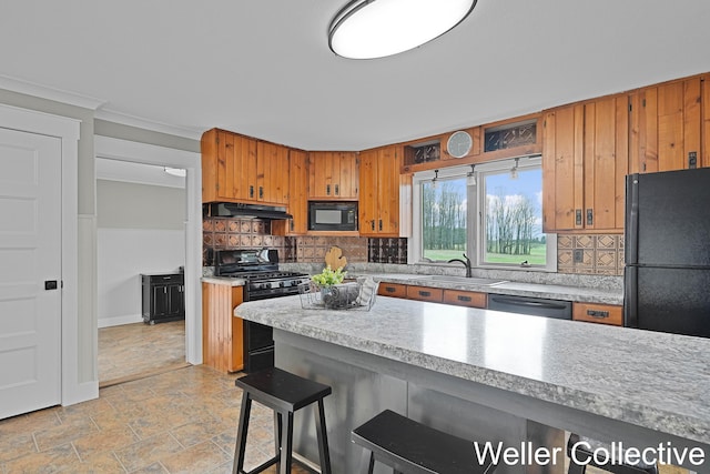 kitchen featuring brown cabinetry, a sink, light countertops, black appliances, and under cabinet range hood
