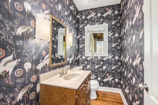 half bath featuring wood finished floors, baseboards, vanity, toilet, and wallpapered walls