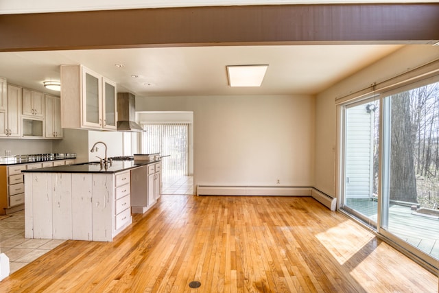 kitchen with wall chimney range hood, dark countertops, a baseboard radiator, glass insert cabinets, and light wood-style flooring