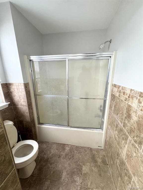 full bath featuring toilet, enclosed tub / shower combo, a wainscoted wall, stone finish flooring, and tile walls
