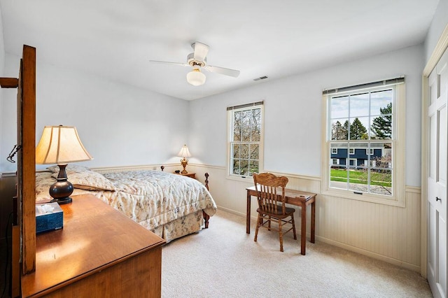 bedroom featuring carpet flooring, a ceiling fan, multiple windows, and a wainscoted wall