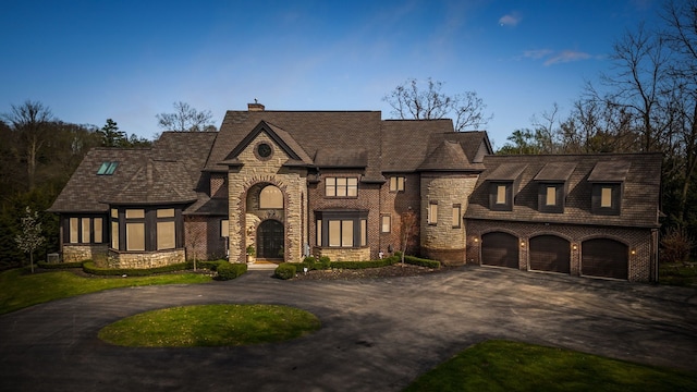 french provincial home featuring an attached garage, brick siding, stone siding, a chimney, and curved driveway