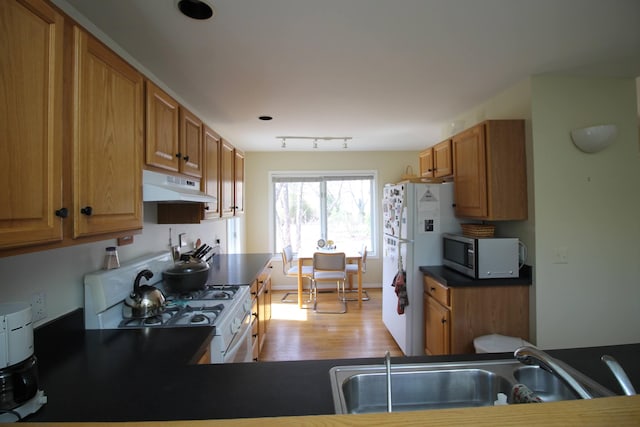 kitchen with dark countertops, white appliances, light wood-type flooring, under cabinet range hood, and a sink