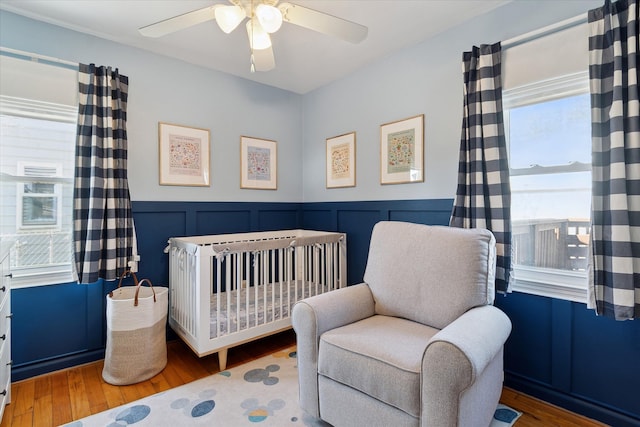 bedroom with a crib, a ceiling fan, wainscoting, and wood finished floors