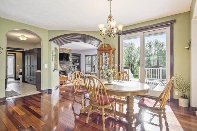 dining area with a notable chandelier, wood-type flooring, arched walkways, a stone fireplace, and baseboards