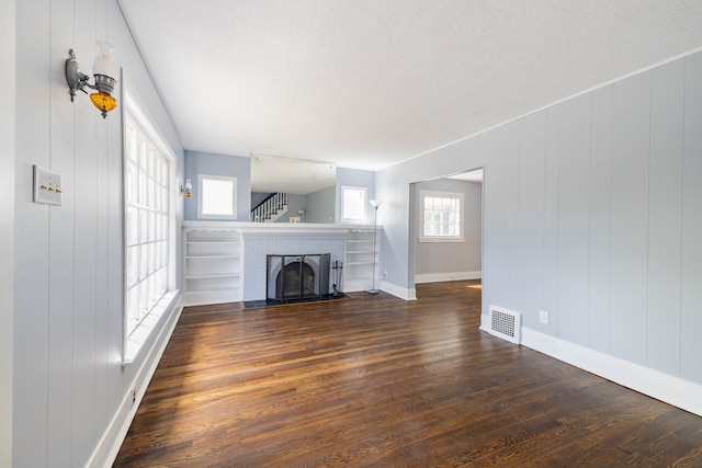 unfurnished living room with a decorative wall, a brick fireplace, baseboards, dark wood-type flooring, and visible vents