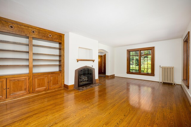 unfurnished living room with arched walkways, a tiled fireplace, radiator heating unit, baseboards, and wood finished floors