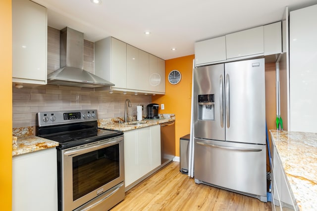kitchen with decorative backsplash, wall chimney range hood, a sink, light wood-style flooring, and appliances with stainless steel finishes