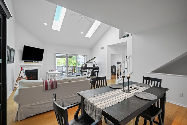 dining room featuring ceiling fan, a skylight, high vaulted ceiling, a warm lit fireplace, and light wood-style flooring