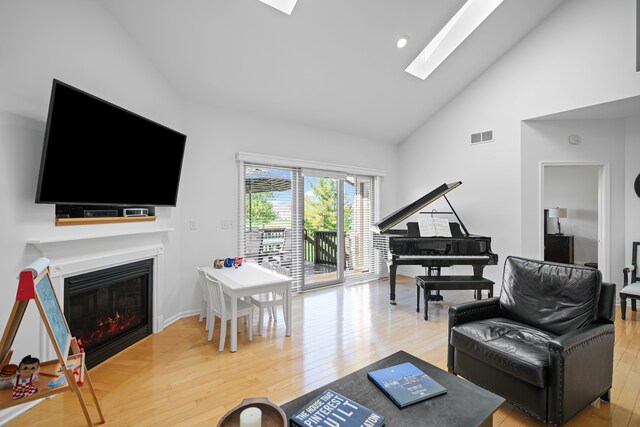 living room with high vaulted ceiling, a glass covered fireplace, wood finished floors, a skylight, and visible vents
